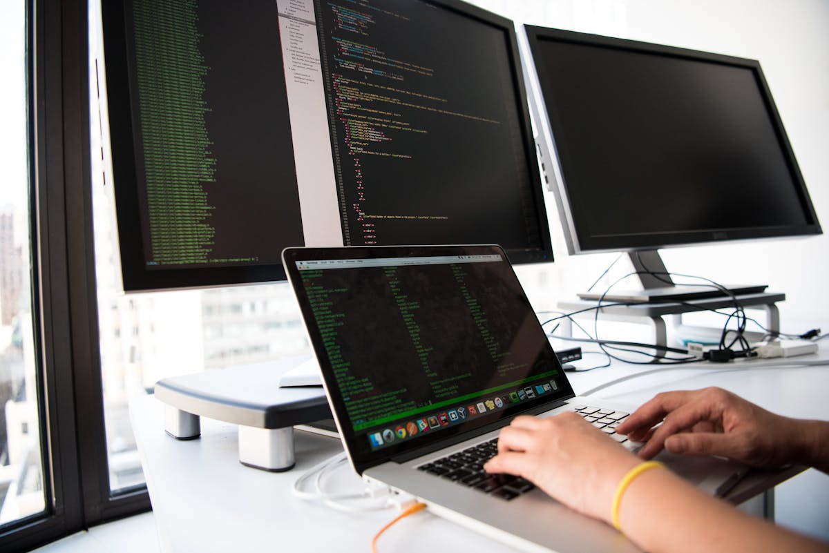 Developer γράφει custom κώδικα σε laptop μπροστά από monitors